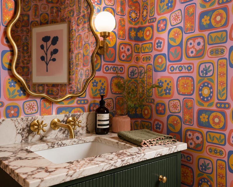 May include: A bathroom with a floral wallpaper in pink, orange, yellow, and blue. A gold-framed mirror hangs above a marble countertop with a white sink and gold fixtures. A bottle of soap and a green washcloth are on the counter.