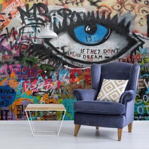 Pode incluir: Uma poltrona azul com uma almofada estampada em branco e dourado fica em frente a uma parede de grafite colorida com um grande olho pintado nela. A parede tamb&eacute;m tem o texto "IF THEY DON'T LET US DREAM THAN WE" escrito nela.