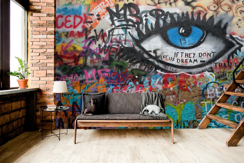 Pode incluir: Um sof&aacute; cinza com uma manta e um animal de pel&uacute;cia fica em frente a uma parede coberta de grafites coloridos. Os grafites apresentam um olho grande com uma &iacute;ris azul e o texto "If they don't let us dream - than we..."