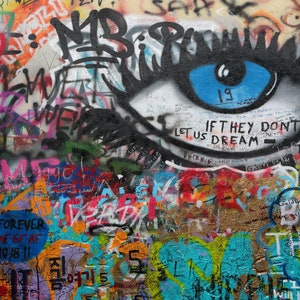 Pode incluir: Uma parede de grafite colorida com um grande olho pintado com uma &iacute;ris azul. O olho &eacute; cercado por v&aacute;rias mensagens e frases, incluindo "If they don't let us dream - than we don't let it the", e "Believe the good of others".