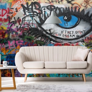 Pode incluir: Um sof&aacute; branco com uma almofada rosa claro fica em frente a uma parede de grafite colorida com um grande olho pintado nela. O olho &eacute; azul com c&iacute;lios pretos e o texto "IF THEY DON'T LET US DREAM - THAN WE DON'T GET THE" est&aacute; escrito abaixo.