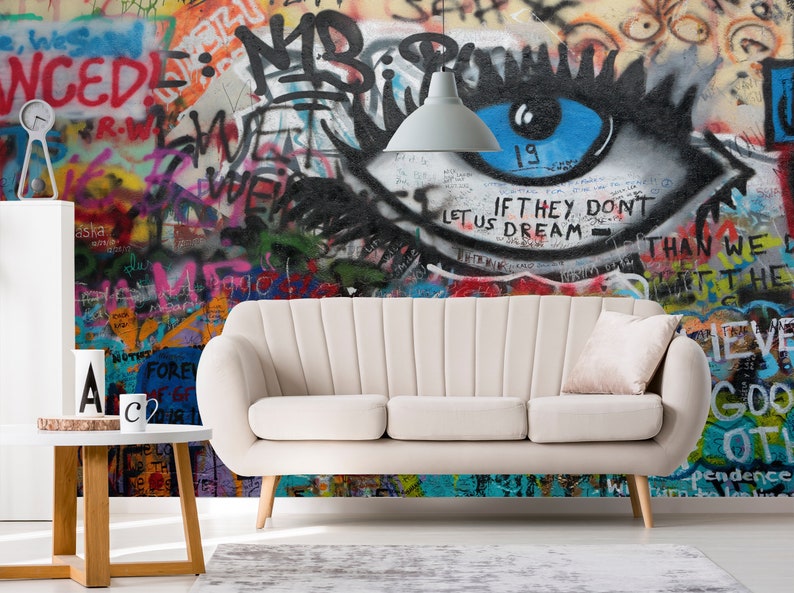 Pode incluir: Um sof&aacute; branco com uma almofada rosa claro fica em frente a uma parede de grafite colorida com um grande olho pintado nela. O olho &eacute; azul com c&iacute;lios pretos e o texto "IF THEY DON'T LET US DREAM - THAN WE DON'T GET THE" est&aacute; escrito abaixo.