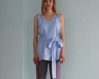 Puede incluir: Una blusa sin mangas de lino azul claro con una cinta para atar. La blusa tiene un escote en V y se lleva con pantalones grises.