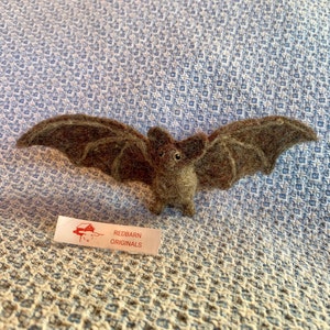 May include: A brown felt bat with black eyes, wings spread out, on a blue and white patterned fabric. A white tag with red lettering reads "Redbarn Originals".