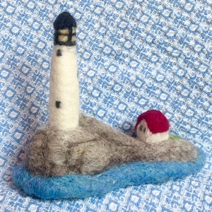 May include: A handcrafted felted wool miniature of a lighthouse on a rocky island. The lighthouse is white with a black top and small windows. A small red-roofed house sits nearby. The island is surrounded by blue felt representing the sea.