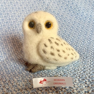 May include: A white and gray needle felted snowy owl with yellow eyes. The owl is sitting on a blue and white patterned fabric. The fabric has a tag that says "Redbarn Originals".