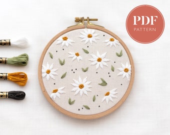May include: A white embroidery hoop with a design of white daisies, green leaves, and black dots on a beige fabric. The hoop is surrounded by four spools of embroidery floss in white, green, orange, and black.