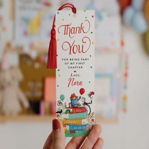 May include: A rectangular paper bookmark with a red tassel at the top. The white surface features colorful confetti dots and the text: Thank You FOR BEING PART OF MY FIRST CHAPTER Love, Nora. Below, illustrations of storybook characters and books.