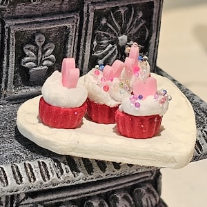 May include: Miniature cupcakes with red bases and white frosting, topped with pink accents and colorful sprinkles, sit on a small, off-white plate. The cupcakes are arranged in front of a decorative, gray, ornate structure.
