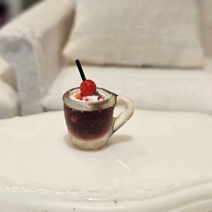 May include: Miniature cocktail in a clear mug with a handle. The drink has a dark red liquid, topped with white foam, sprinkles, a red cherry, and a black straw. The mug is sitting on a white surface.