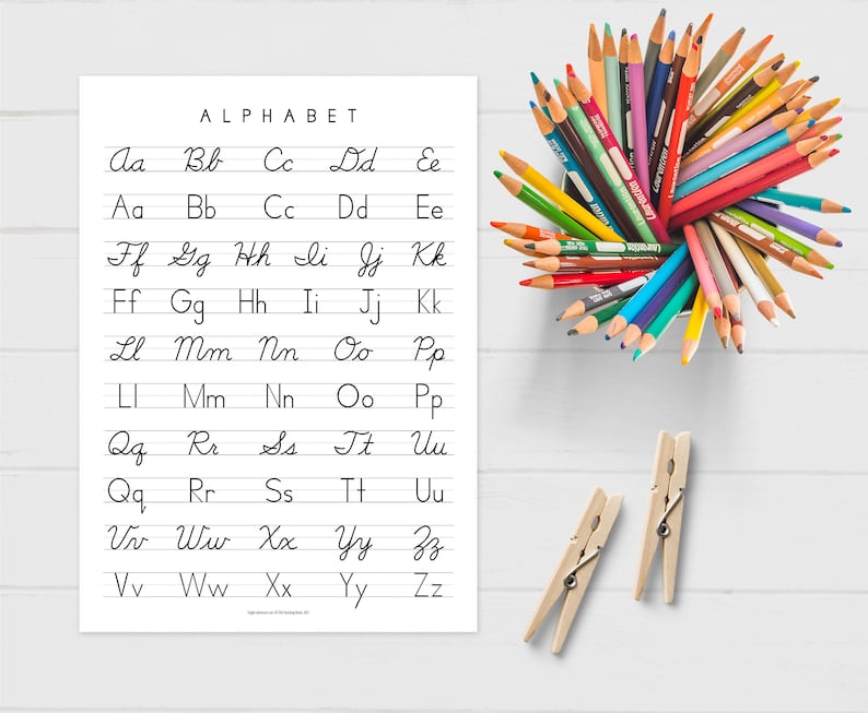 May include: A printable worksheet with the alphabet written in cursive. The letters are written in black ink on white paper with lined paper. There are two lines for each letter. There are also colored pencils in a cup and two wooden clothespins.