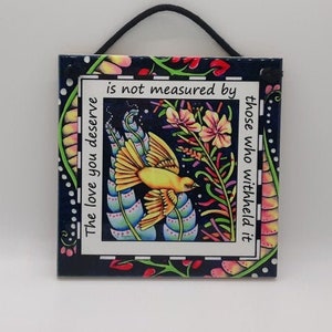 May include: A colorful tile with a blue background and a white border. The tile features a yellow bird with a black eye and a floral design. The text on the tile reads "The love you deserve is not measured by those who withheld it."