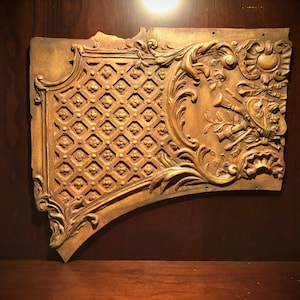 May include: A gold-toned, ornate, wooden wall decor piece with a carved design featuring a floral pattern and a shield with a trumpet motif.