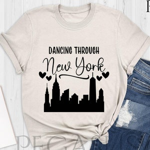 May include: A beige t-shirt with the text "Dancing Through New York" in black lettering with a black silhouette of the New York City skyline and hearts above the skyline. The t-shirt is on a white wooden surface with a pair of blue jeans, a notebook, and purple sneakers.