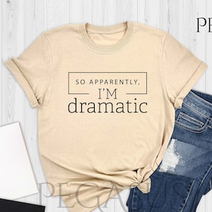 May include: A beige t-shirt with the text "So apparently, I'm dramatic" printed on it. The shirt is laid out on a white wooden surface with a pair of blue jeans, a notebook, and a pair of green sneakers.