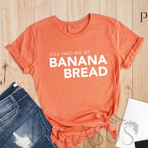 May include: A heather orange t-shirt with the text "You had me at banana bread" printed in white.