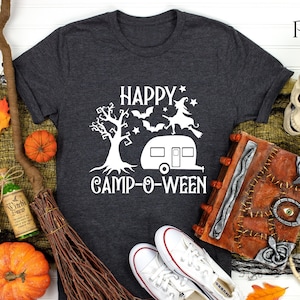 May include: Dark grey t-shirt with a white graphic reading "HAPPY CAMP-O-WEEN", featuring a tree, witch, bats, and a camper. The image includes Halloween decorations such as pumpkins, a broom, and a skull.