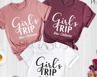 May include: Three white t-shirts with black text that reads "Girl's Trip New Orleans". The shirts are laid out on a light wood surface with a bouquet of pink and yellow flowers, a laptop, a cup of tea, and sunglasses.