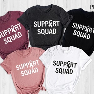 May include: Five t-shirts in different colors with the text "Support Squad" and a white ribbon graphic. The colors are heather maroon, heather dark grey, black, heather mauve, and white.