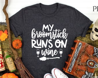 May include: Dark gray t-shirt with the text "MY broomstick RUNS ON wine" in white. The image includes Halloween decorations like pumpkins, a broom, a skull, and a spell book. White sneakers are also present.