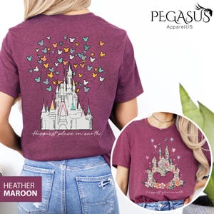 Op de afbeelding: Een heide-maron t-shirt met een afbeelding van Assepoester's kasteel en Mickey Mouse ballonnen. De tekst "Happiest place on earth" is onder het kasteel gedrukt. De achterkant van het shirt heeft een vergelijkbaar ontwerp met een bloemenpatroon.