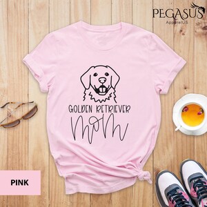 Op de afbeelding: Roze T-shirt met een zwarte omtrek van een golden retriever en de tekst "Golden Retriever Mom".