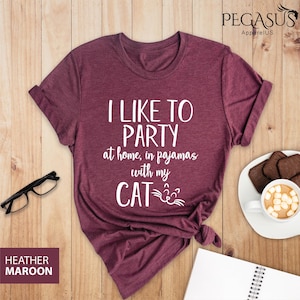 Op de afbeelding: Heather-marron T-shirt met de tekst "Ik vind het leuk om thuis in mijn pyjama te feesten met mijn kat" in witte letters. De kat heeft een wit gezicht met een zwarte neus en snorharen.