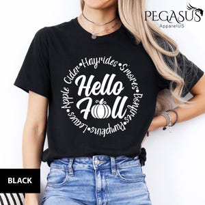 May include: Black t-shirt with a white circular design that says "Hello Fall" with a pumpkin graphic. The design also includes the words "Cider, Hayrides, S'mores, Bonfires, Pumpkins, Leaves, and Apple."
