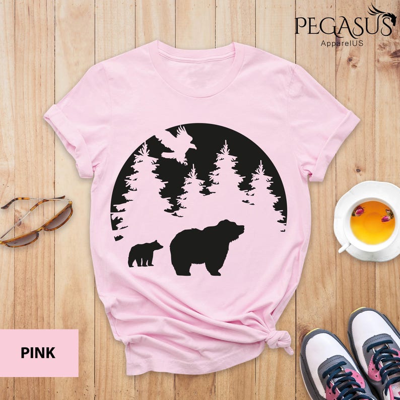 Puede incluir: Camiseta rosa con una silueta negra de un oso y un cachorro en una escena de bosque. El dise&ntilde;o est&aacute; dentro de un c&iacute;rculo con &aacute;rboles y un p&aacute;jaro volando por encima. El texto "PEGASUS ApparelUS" est&aacute; en la esquina superior derecha.
