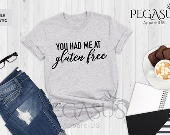 May include: A heather grey t-shirt with the text "You had me at gluten free" printed in black. The shirt is laid flat on a white wooden surface with a pair of jeans, glasses, a notebook, and a cup of hot chocolate with marshmallows.