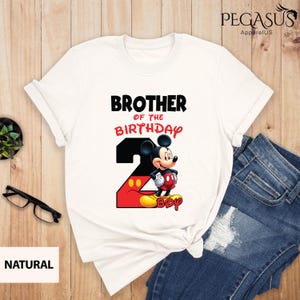 May include: White t-shirt featuring the text "BROTHER OF THE BIRTHDAY" above a graphic of Mickey Mouse and the number 2. The number 2 is red and black with yellow accents. The word "BOY" is below Mickey Mouse.