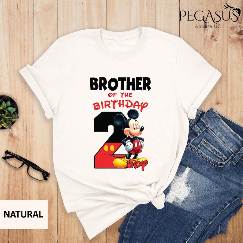 May include: White t-shirt featuring the text "BROTHER OF THE BIRTHDAY" above a graphic of Mickey Mouse and the number 2. The number 2 is red and black with yellow accents. The word "BOY" is below Mickey Mouse.