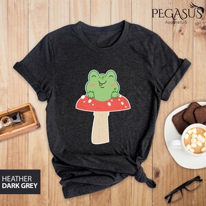 May include: Dark heather grey t-shirt with a cartoon frog sitting on a red and white mushroom. The frog is green with a big smile and a long tongue sticking out.