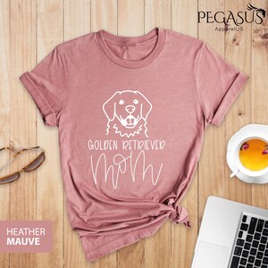 Op de afbeelding: Een heide-paarse t-shirt met een witte omtrek van een golden retriever hond en de tekst "Golden Retriever Mom".