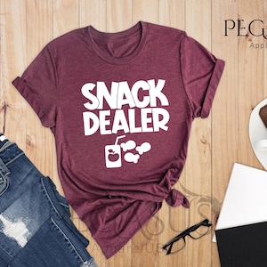 Peut inclure: Un t-shirt en marron chin&eacute; avec le texte "Snack Dealer" en blanc. Le t-shirt est nou&eacute; en bas et comporte un graphique d'une bo&icirc;te de jus avec une pomme et du pop-corn.