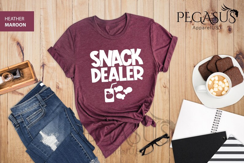 Peut inclure: Un t-shirt en marron chin&eacute; avec le texte "Snack Dealer" en blanc. Le t-shirt est nou&eacute; en bas et comporte un graphique d'une bo&icirc;te de jus avec une pomme et du pop-corn.