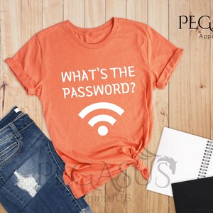 May include: A heather orange t-shirt with the text "What's the password?" and a white wifi symbol. The shirt is on a wooden surface with a pair of jeans, a notebook, and a plant.