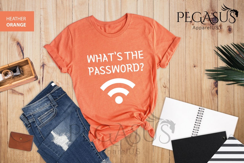 May include: A heather orange t-shirt with the text "What's the password?" and a white wifi symbol. The shirt is on a wooden surface with a pair of jeans, a notebook, and a plant.