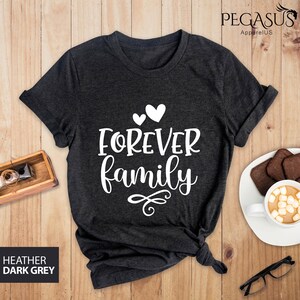 Pode incluir: Uma camiseta cinza escuro com o texto "FOREVER family" em letras brancas. Dois pequenos cora&ccedil;&otilde;es brancos est&atilde;o acima do texto. A camiseta est&aacute; colocada sobre uma superf&iacute;cie de madeira.