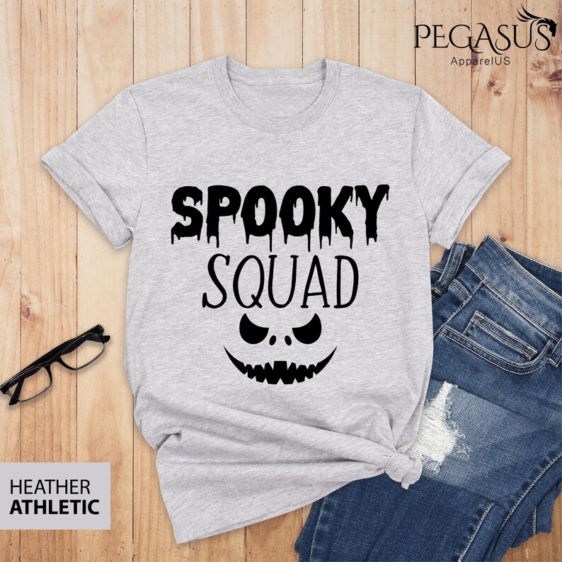 May include: Heather athletic gray t-shirt with the words "SPOOKY SQUAD" in black, featuring a jack-o'-lantern face design. The shirt is paired with blue jeans and black glasses. The brand name "Pegasus ApparelUS" is visible.