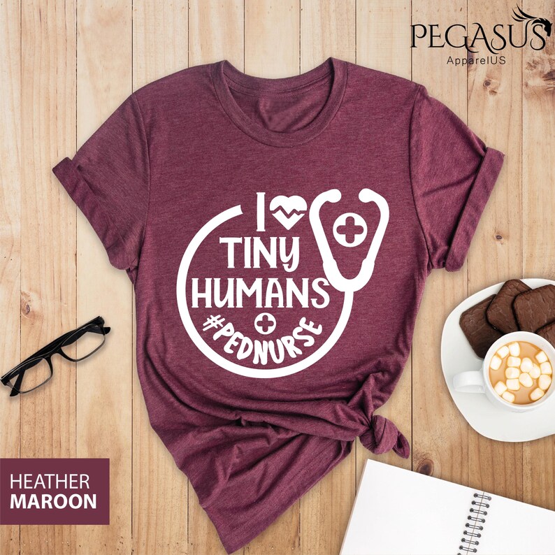 Op de afbeelding: Een maroon t-shirt met een witte afbeelding waarop staat "I love tiny humans #pednurse" met een stethoscoop en een hartontwerp.