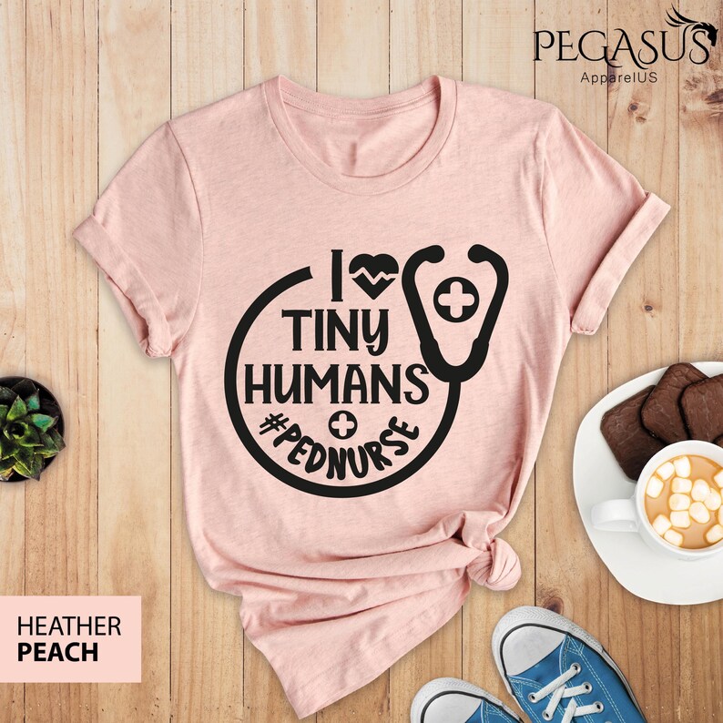 Op de afbeelding: Een roze T-shirt met een zwarte afbeelding waarop staat "Ik hou van kleine mensen #pednurse" met een stethoscoop en een hart.