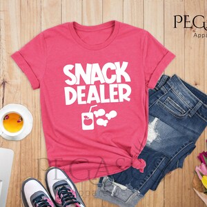 Peut inclure: Un t-shirt rose avec le texte "Snack Dealer" en blanc. Le t-shirt a un graphique d'une bo&icirc;te de jus avec une pomme et des poissons.