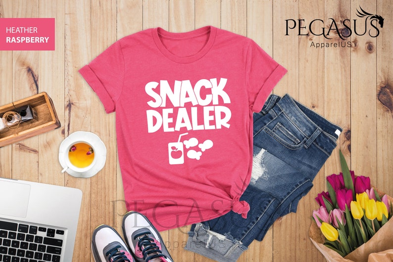 Peut inclure: Un t-shirt rose avec le texte "Snack Dealer" en blanc. Le t-shirt a un graphique d'une bo&icirc;te de jus avec une pomme et des poissons.