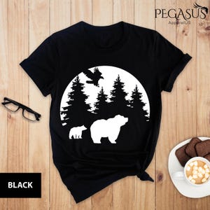 Puede incluir: Camiseta negra con una silueta blanca de un oso, dos osos m&aacute;s peque&ntilde;os y un p&aacute;jaro volando sobre una luna con &aacute;rboles detr&aacute;s.