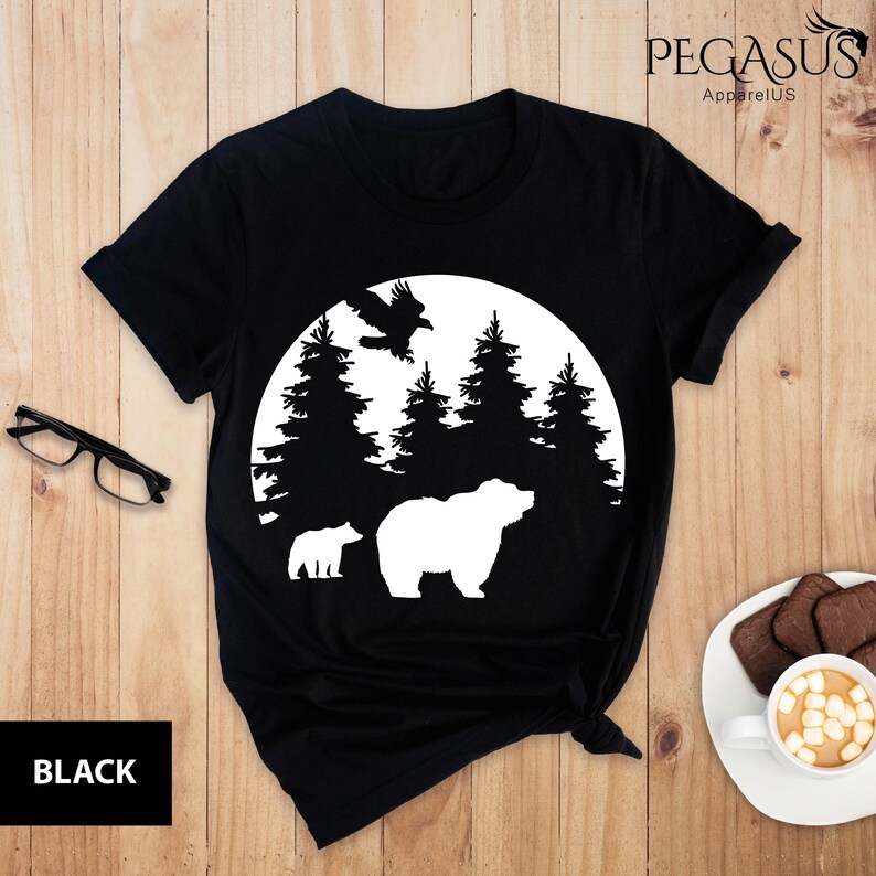 Puede incluir: Camiseta negra con una silueta blanca de un oso, dos osos m&aacute;s peque&ntilde;os y un p&aacute;jaro volando sobre una luna con &aacute;rboles detr&aacute;s.