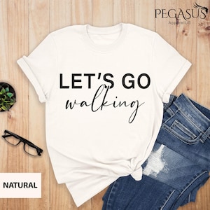 May include: White t-shirt with black text that reads "Let's Go Walking" on a white background. The shirt is laid out on a wooden surface with a pair of blue jeans and a plant.