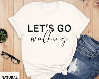 May include: White t-shirt with black text that reads "Let's Go Walking" on a white background. The shirt is laid out on a wooden surface with a pair of blue jeans and a plant.