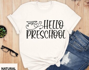May include: White t-shirt with black text that says "Hello Preschool" with a paper airplane and hearts graphic. The shirt is on a wooden surface with a pair of jeans.