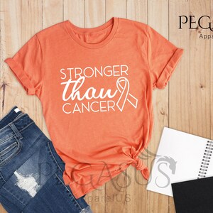 May include: Heather orange t-shirt with the text "Stronger than Cancer" in white with a white ribbon graphic.  The shirt is laid flat on a wood surface.